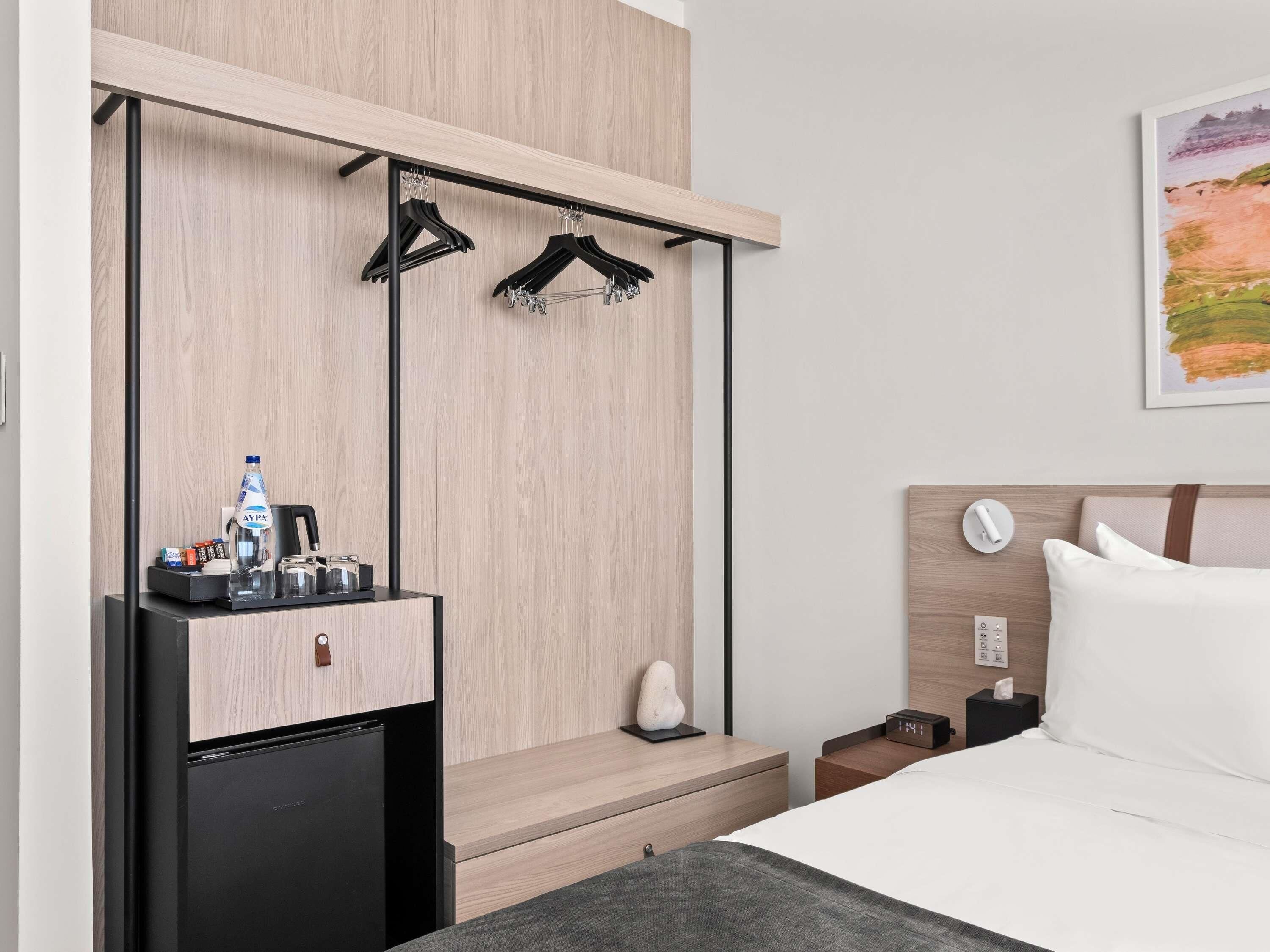 Une chambre d’hôtel comprenant un lit, un petit réfrigérateur, une table de chevet avec une lampe, une étagère murale avec des cintres et un tableau au mur.