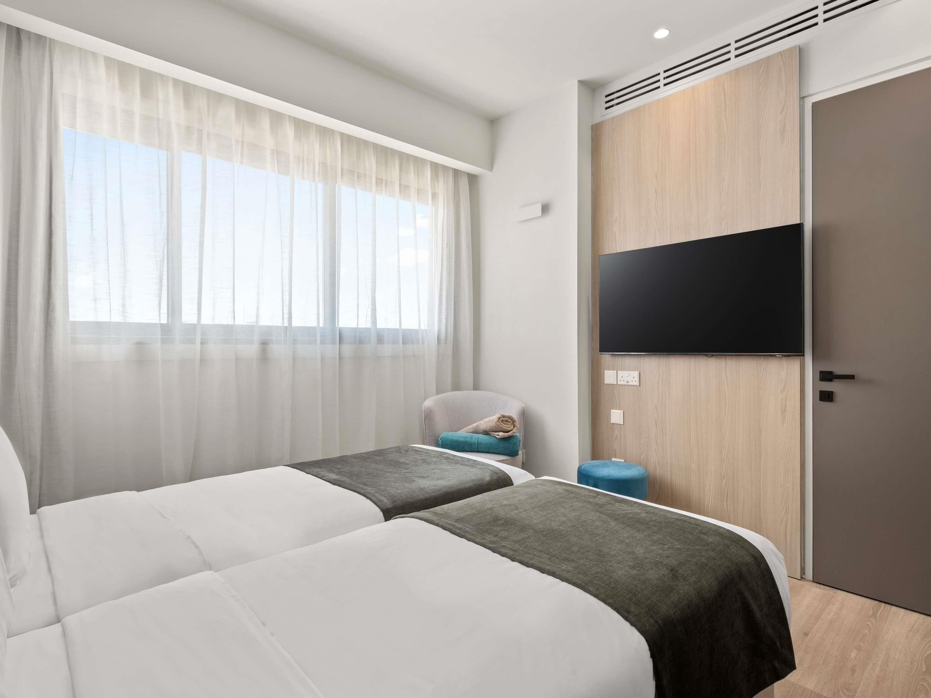 A hotel room with two beds, a TV mounted on the wall, a window with sheer curtains, and a wooden wardrobe.