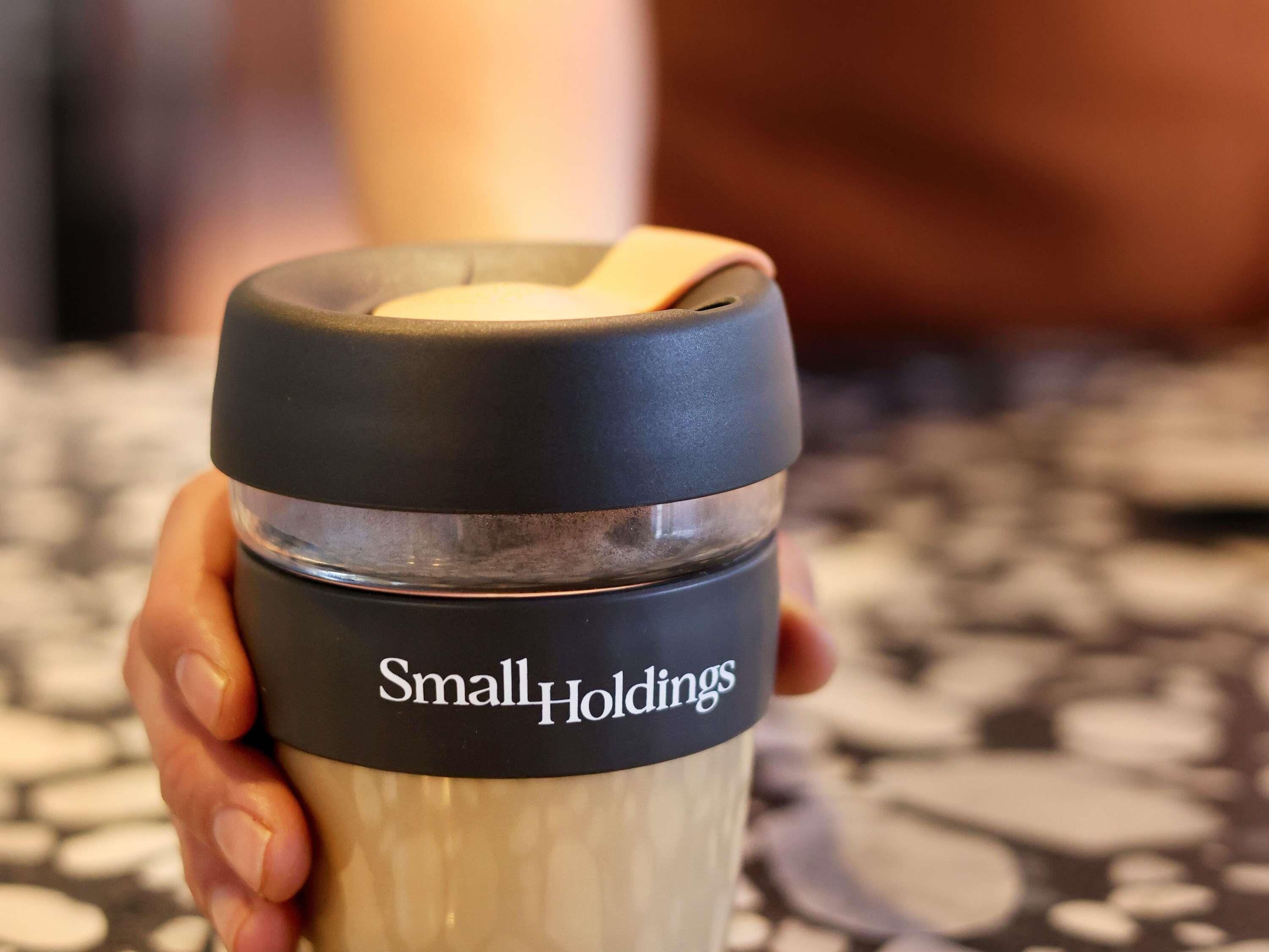 Une main tenant une bouteille d’eau avec un bouchon noir et une étiquette sur laquelle on peut lire « SmallHoldings ».