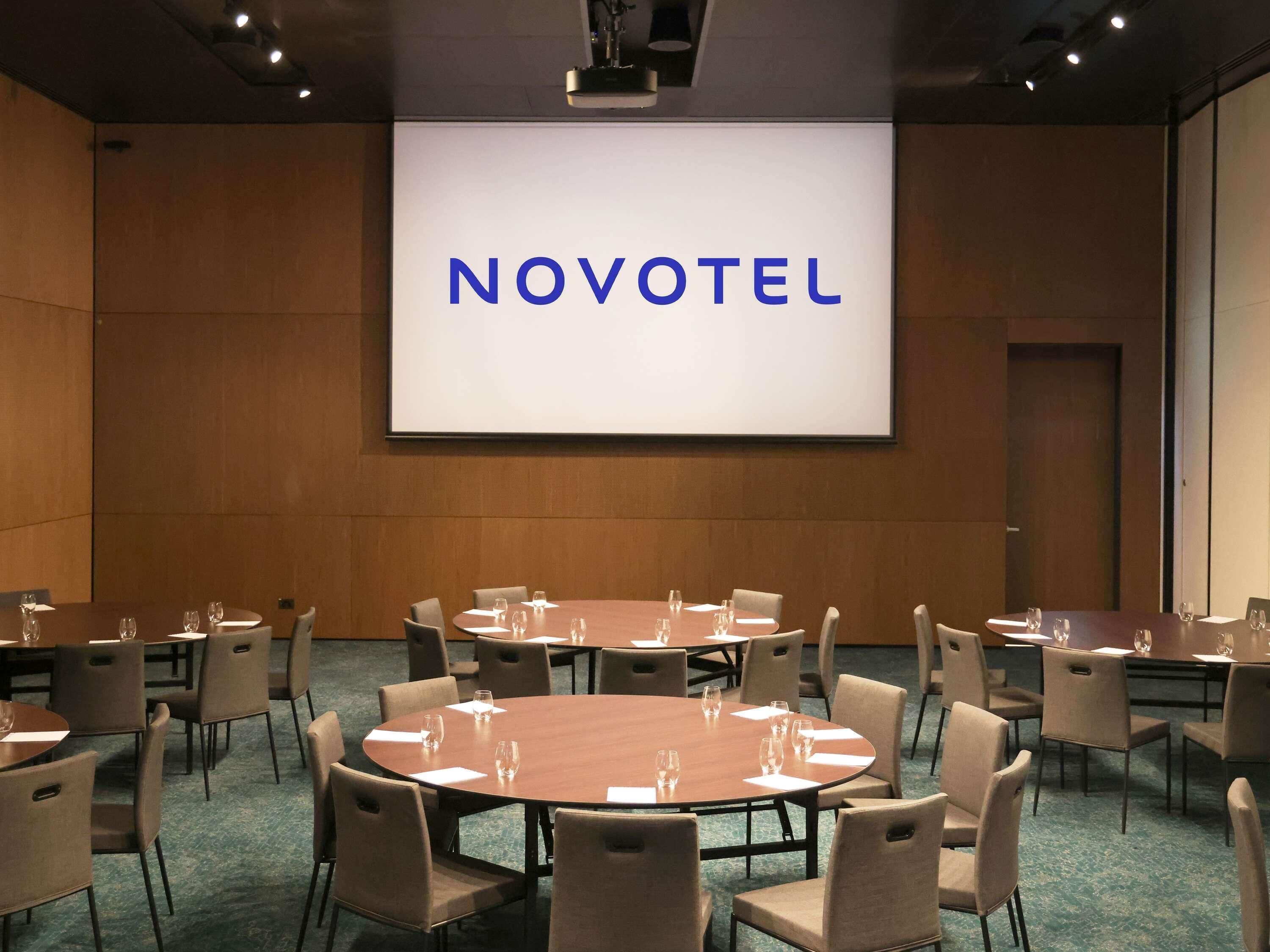 Une salle de conférence avec plusieurs tables et chaises, un grand écran affichant le mot « NOVOTEL », et un mur en bois.