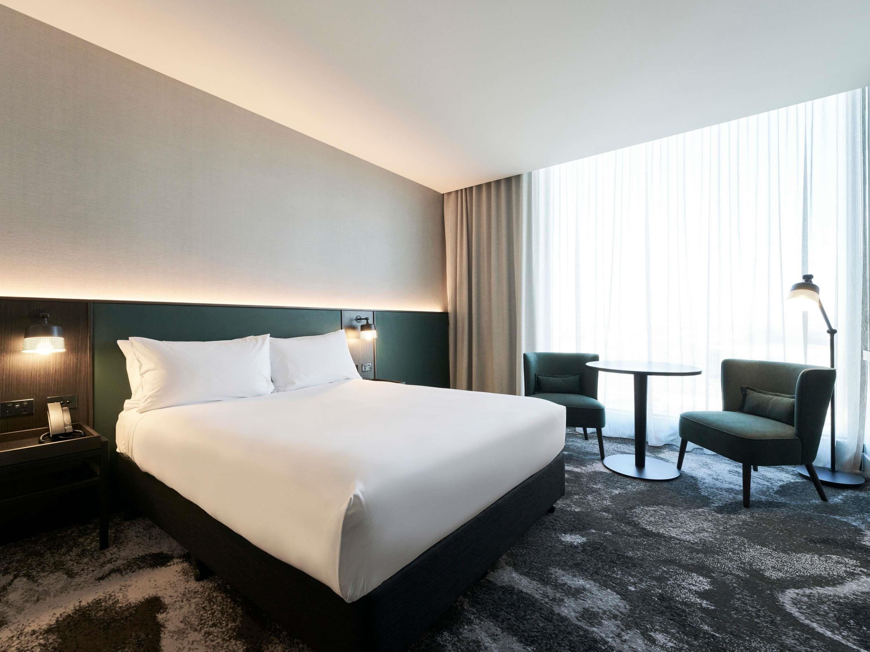 Habitación de hotel con una cama grande, dos lámparas de noche, una mesita pequeña, dos sillas y un ventanal con cortinas.