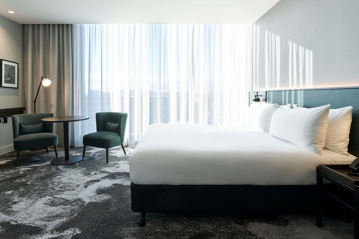 Ein Hotelzimmer mit einem großen Bett, zwei Stühlen, einem kleinen Tisch und Blick auf die Stadt durch transparente Vorhänge.