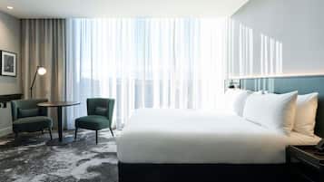 A hotel room with a large bed, two chairs, a small table, and a view of the city through sheer curtains.