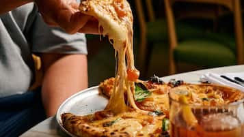 A person holding a slice of pizza with melted cheese, a glass of a dark beverage, and a plate with remnants of pizza on a table.