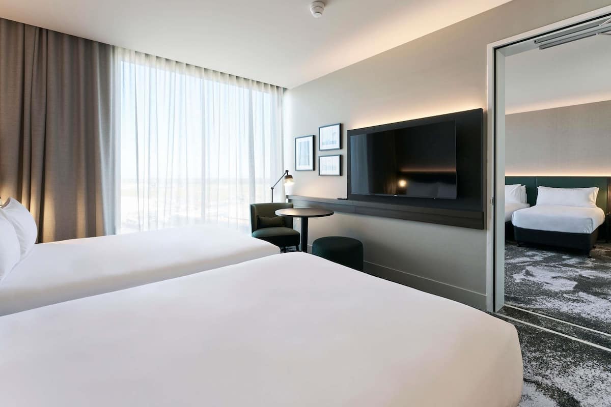 Ein Hotelzimmer mit zwei Betten, einem Fernseher, einem Schreibtisch und einem großen Fenster mit Vorhängen.
