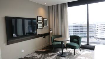 A hotel room with a large TV, a small round table, two green chairs, a lamp, and a view of the city through large windows.