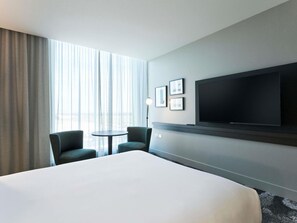 Une chambre d’hôtel avec un grand lit, deux chaises, une petite table, un téléviseur fixé au mur et une fenêtre avec des rideaux.