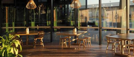 An indoor dining area with wooden tables and chairs, large windows, and hanging pendant lights.