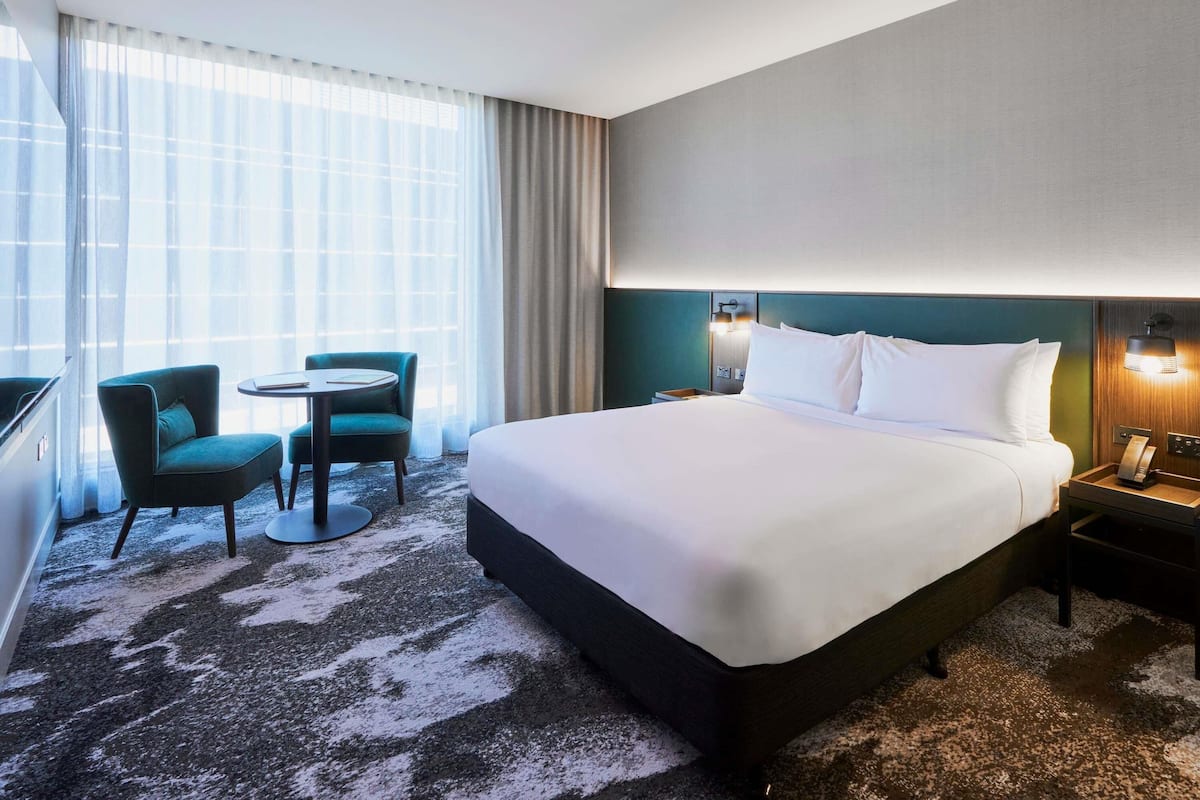 Ein Hotelzimmer mit einem großen Bett, einem Schreibtisch, zwei Stühlen, einem kleinen Tisch und einem großen Fenster mit Vorhängen.