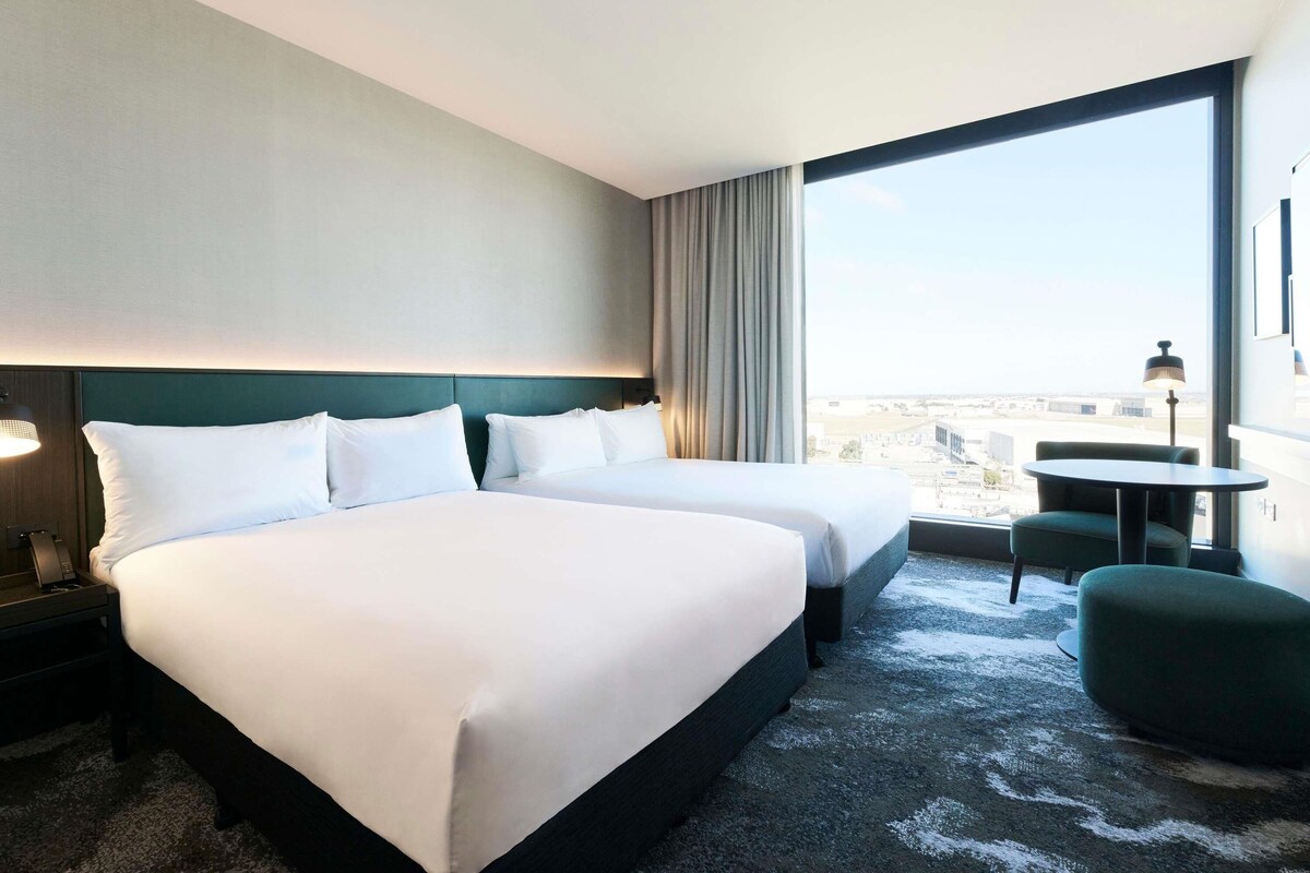 Habitación de hotel con dos camas, vistas a la ciudad, una mesita y una silla.