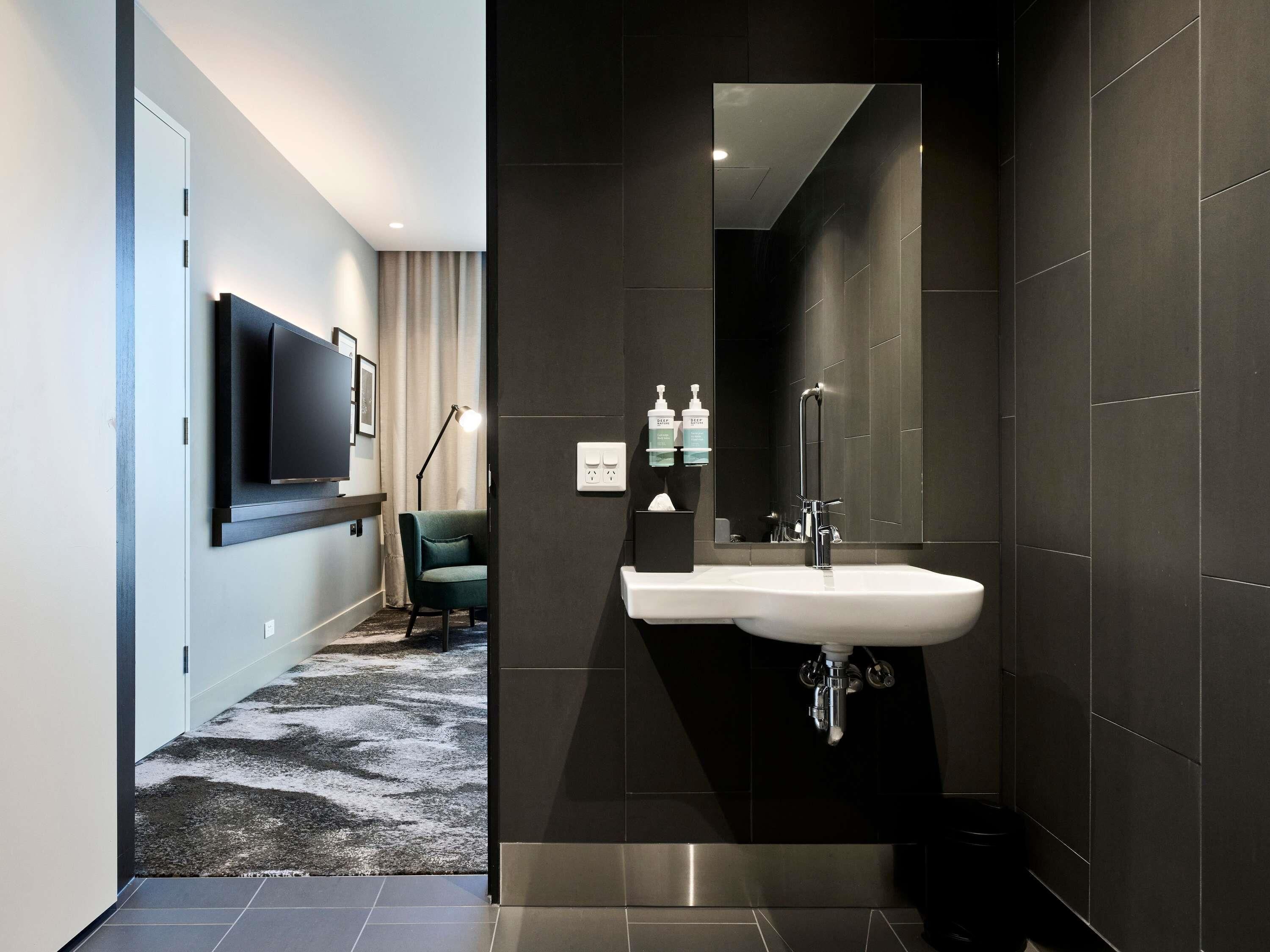 Un baño moderno con un espejo grande, baldosas oscuras, un lavamanos blanco y un cubo de basura negro.