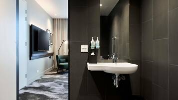 Un baño moderno con un espejo grande, baldosas oscuras, un lavamanos blanco y un cubo de basura negro.