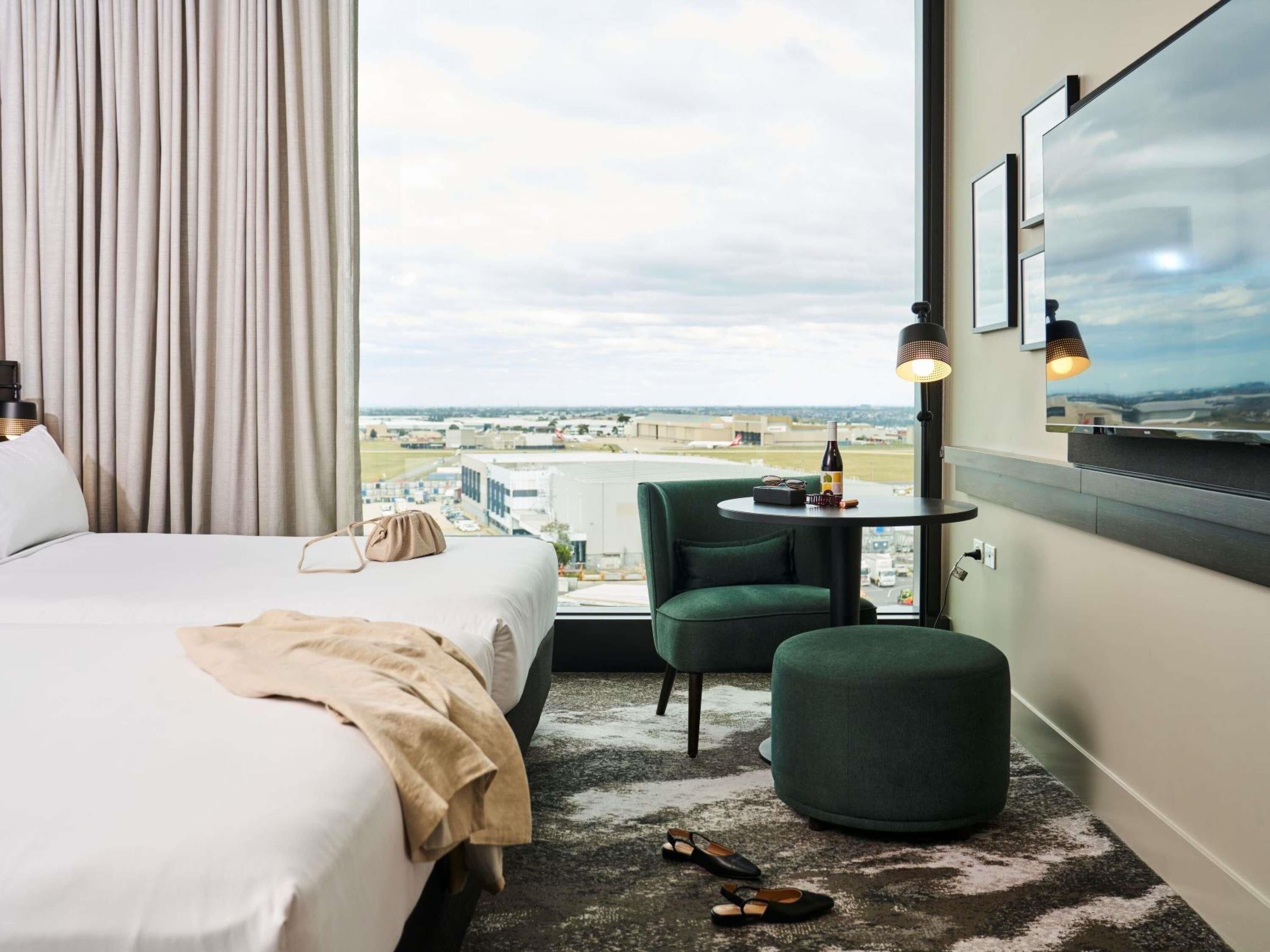 Habitación de hotel con una cama, un sillón verde, una mesa redonda con una botella y vasos, y vista al aeropuerto.