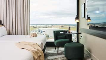 Habitación de hotel con una cama, un sillón verde, una mesa redonda con una botella y vasos, y vista al aeropuerto.