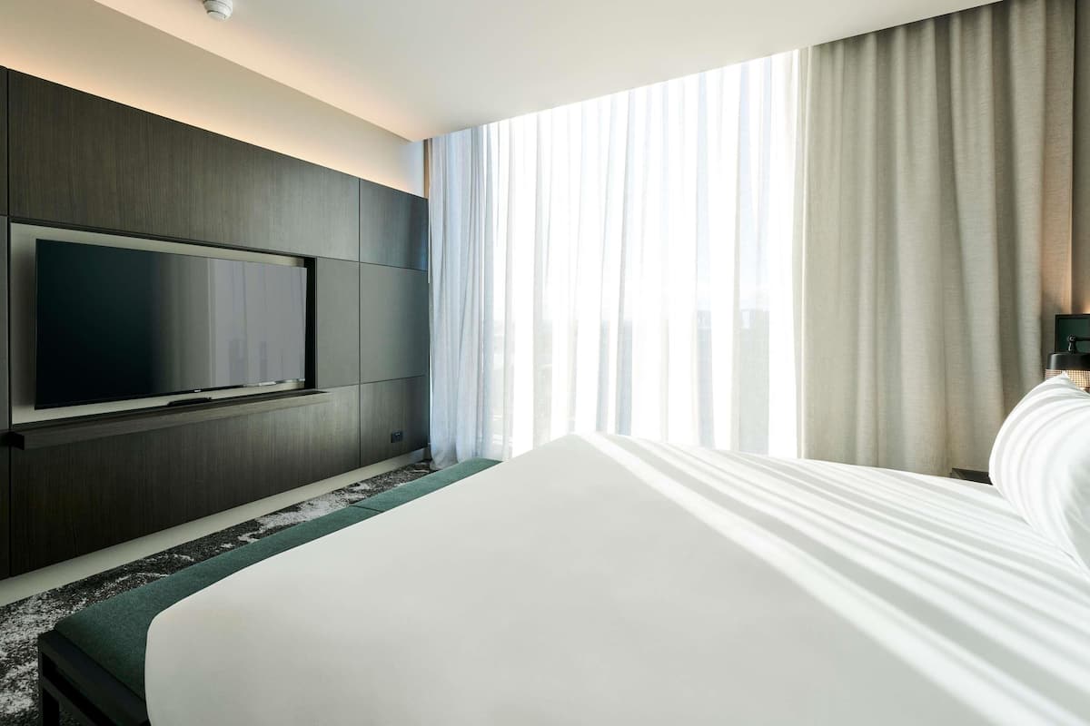 Ein Hotelzimmer mit einem großen Bett, einem an der Wand befestigten Flachbildfernseher und einem Fenster mit durchsichtigen Vorhängen.
