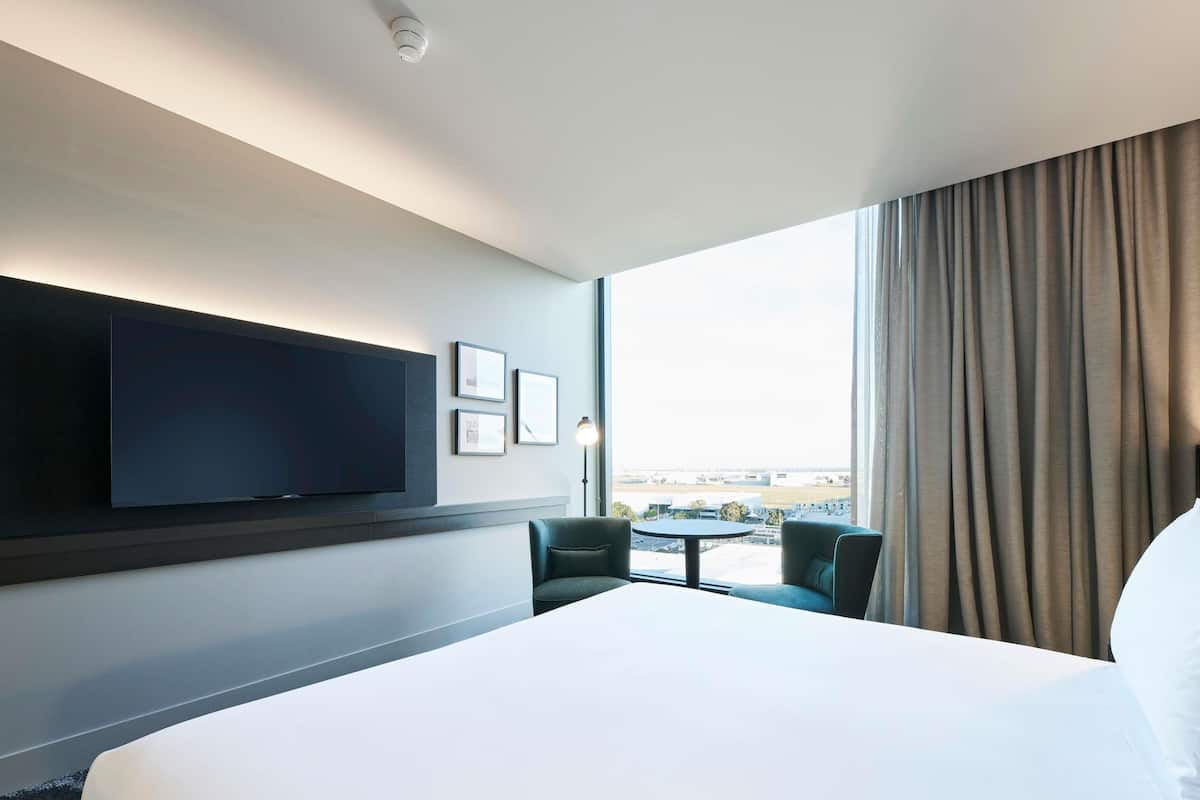 Ein Hotelzimmer mit einem großen Bett, einem an der Wand befestigten Flachbildfernseher, einem kleinen Tisch, zwei Stühlen und einem Fenster mit Vorhängen.