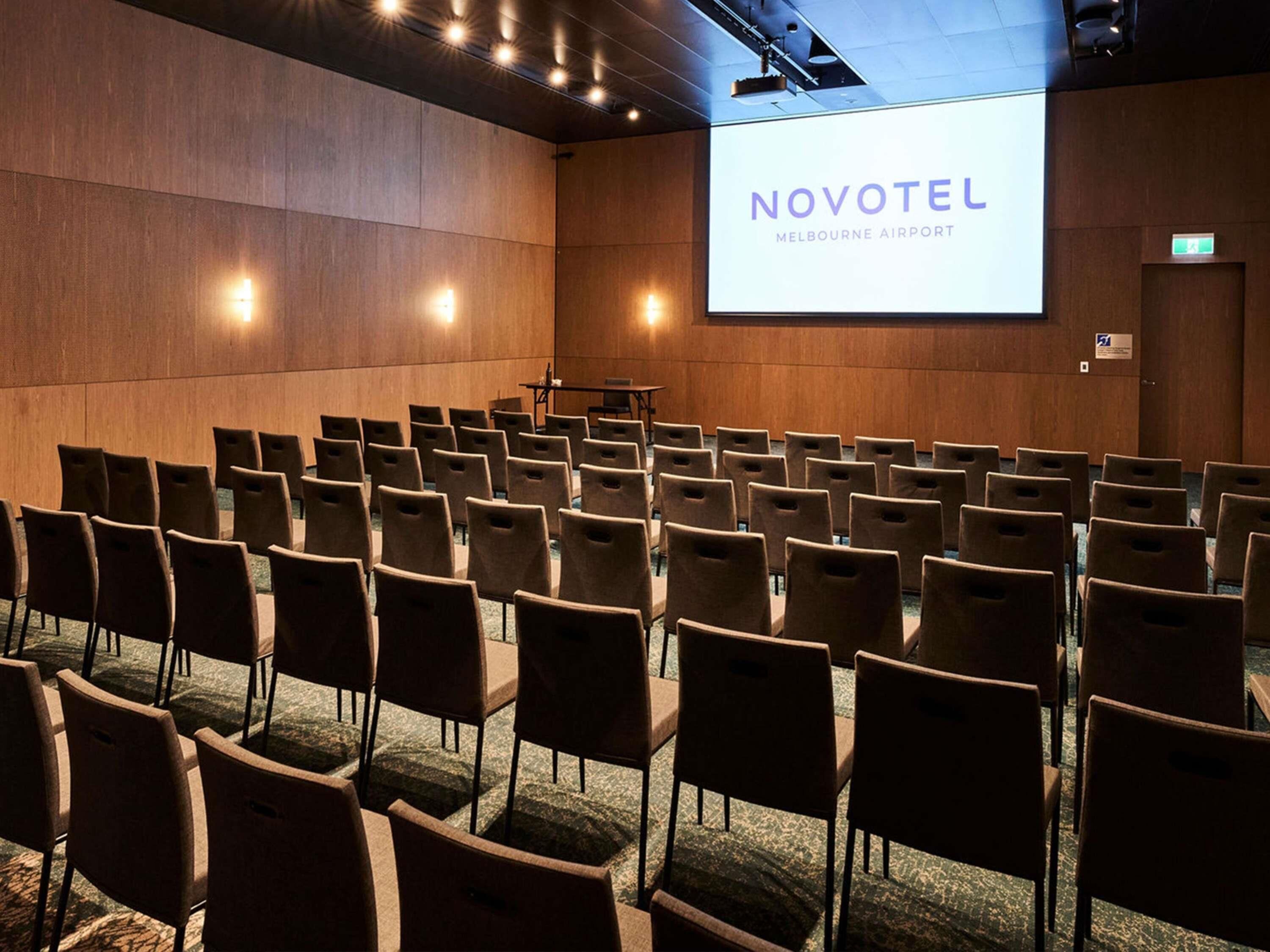 Une salle de conférence avec des rangées de chaises, un pupitre et un écran affichant la mention « NOVOTEL MELBOURNE AIRPORT ».