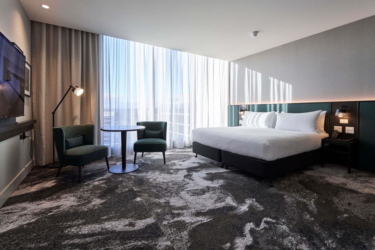A hotel room with a large bed, a desk, two chairs, a TV, and a view of the city through sheer curtains.