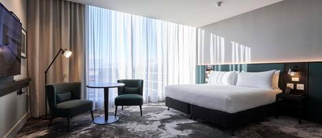 Habitación de hotel con una cama grande, un escritorio, dos sillas, un televisor y vistas de la ciudad a través de cortinas translúcidas.