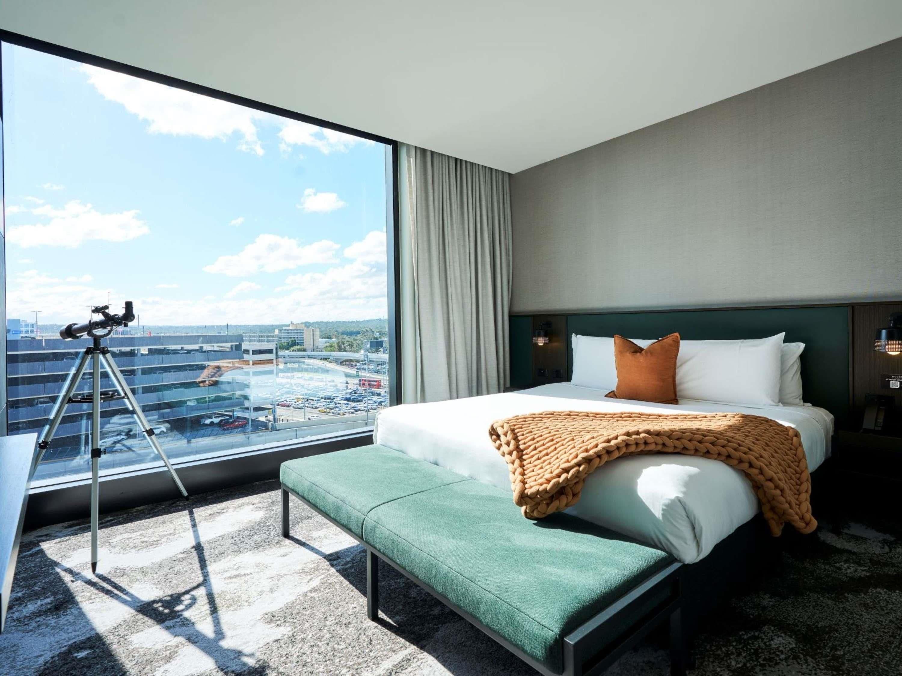 A hotel room with a large window, a telescope, a bed with pillows, a bench, and a view of a parking garage.