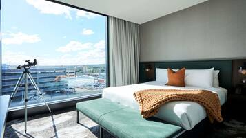 A hotel room with a large window, a telescope, a bed with pillows, a bench, and a view of a parking garage.