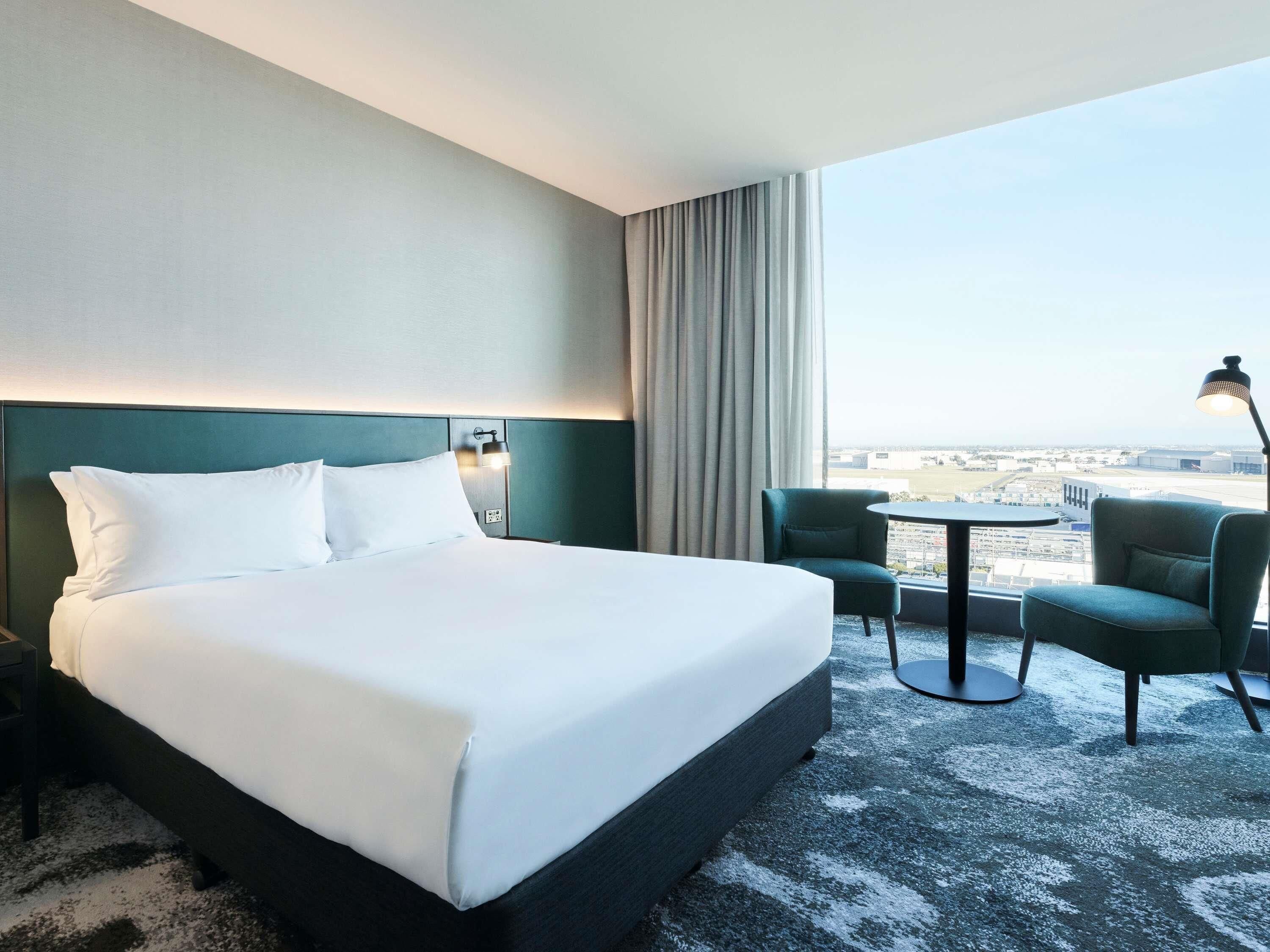 Camera d'albergo con un letto grande, due sedie, un tavolino e vista sull'aeroporto.