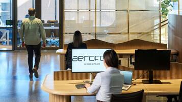 A modern workspace with a sign reading 'aeroFOC WORKSPACE,' featuring a curved wooden desk, a laptop, and a person working.