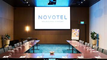 Una sala de conferencias con una pantalla grande que muestra 'NOVOTEL MELBOURNE AIRPORT'.