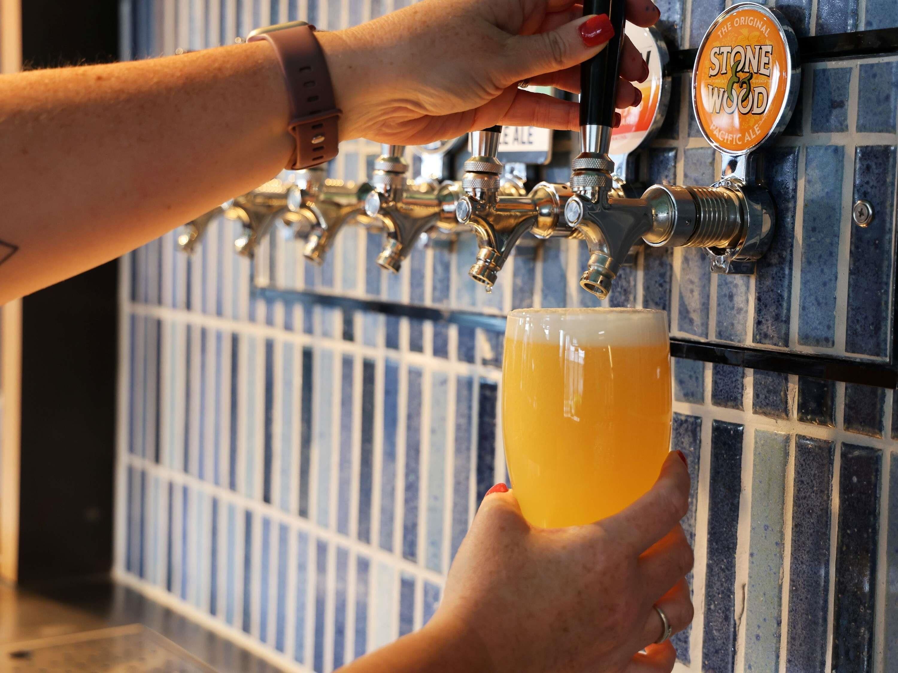 A hand holding a beer mug under a tap labeled 'Stone Wood Pacific Ale'.
