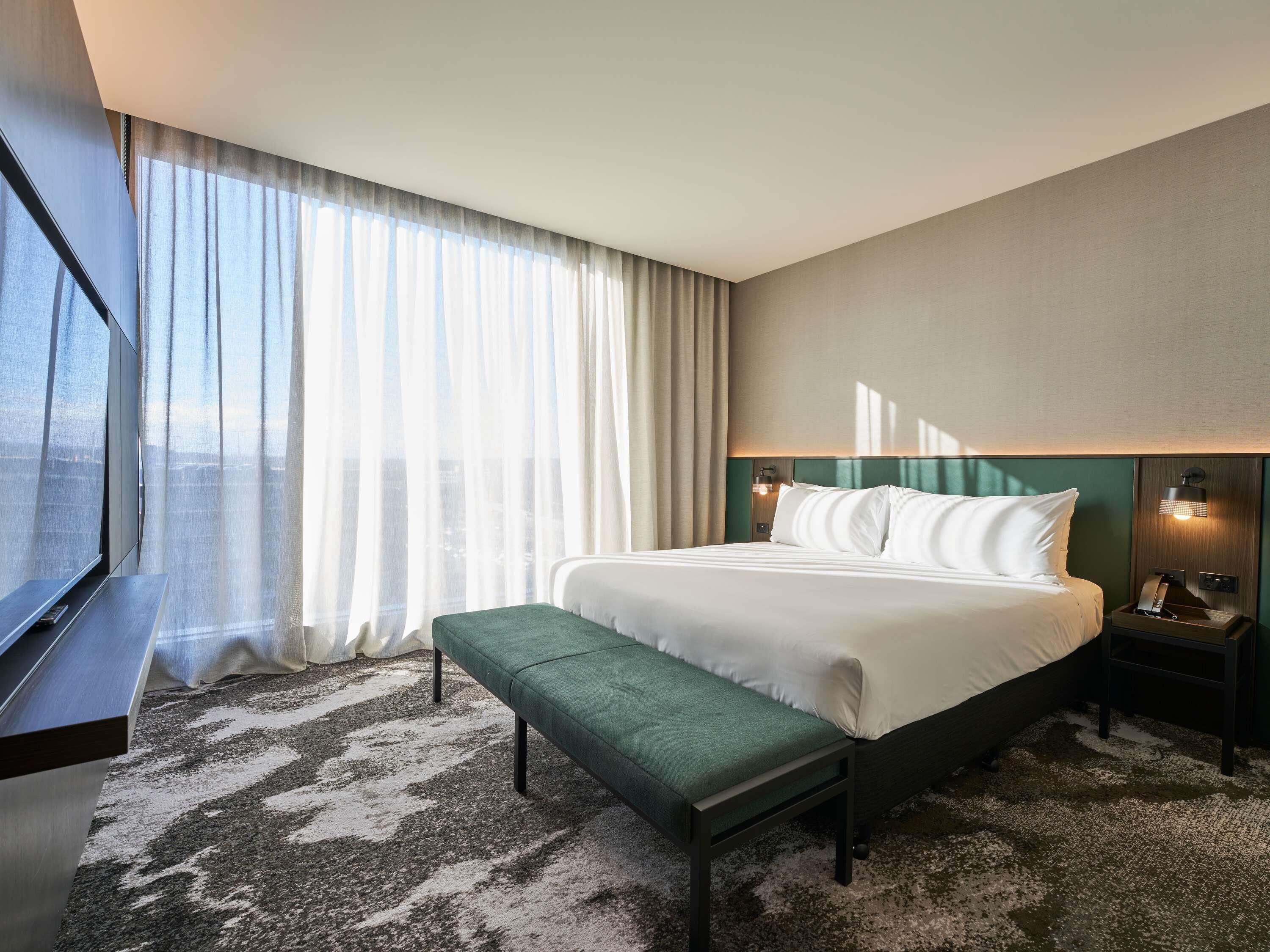 Habitación de hotel con cama, banco, mesita de noche, lámpara y ventana con cortinas.