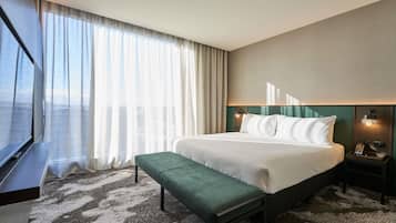 Habitación de hotel con cama, banco, mesita de noche, lámpara y ventana con cortinas.