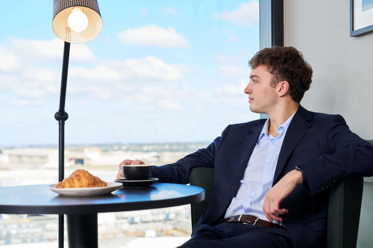 A person seated at a table with a croissant and a cup, near a lamp with a city view.