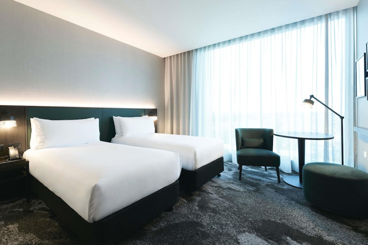 Ein Hotelzimmer mit zwei Betten, einem Schreibtisch, einem Stuhl und einem großen Fenster mit Vorhängen.