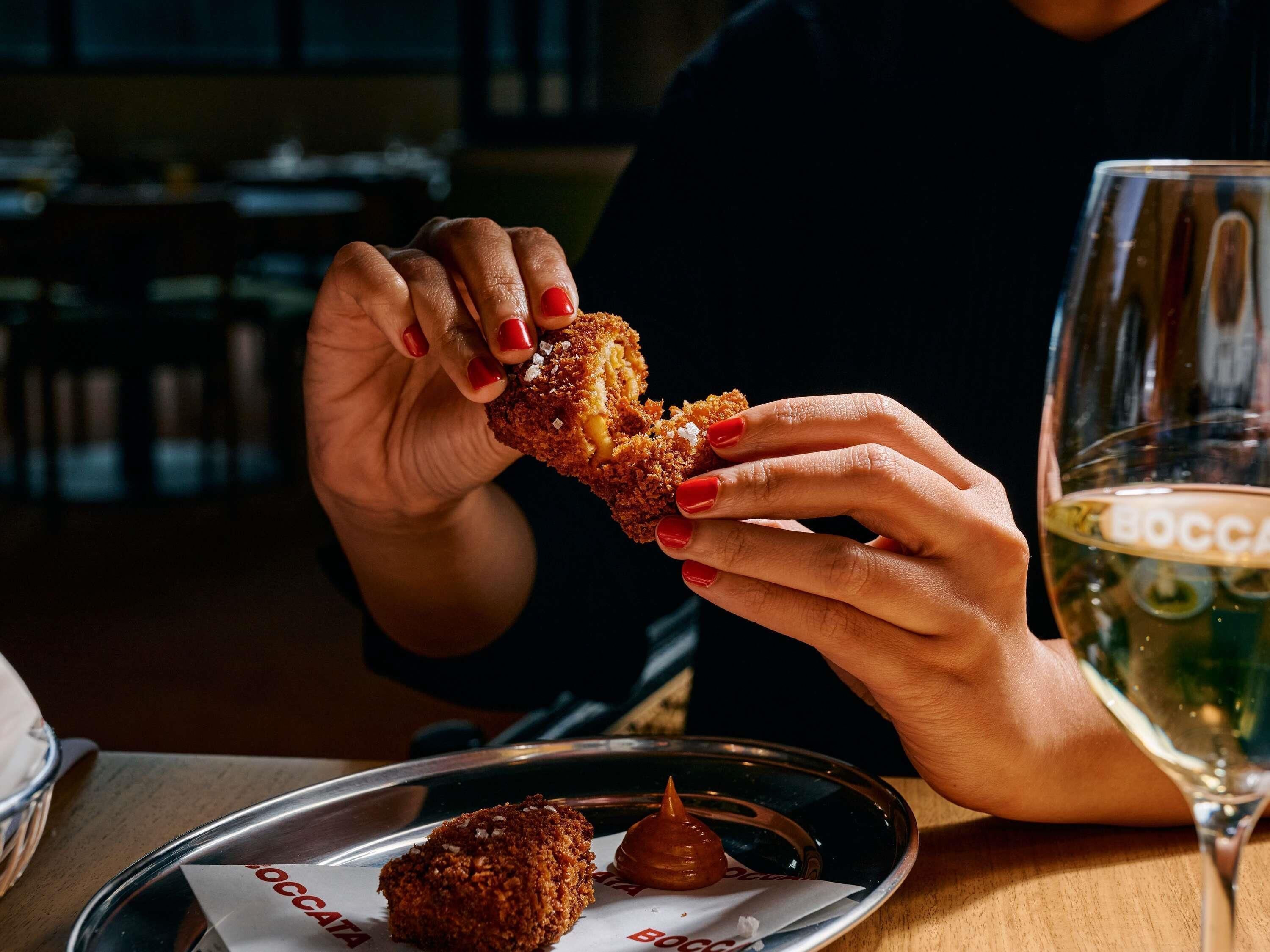 En person håller i en panerad maträtt med sås på en tallrik, och bredvid står ett glas vitt vin.