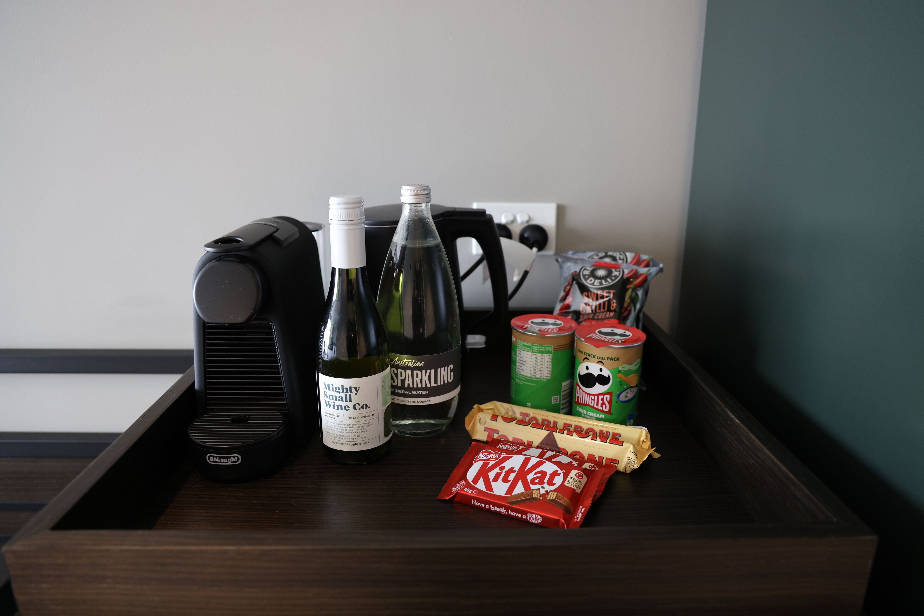 A wooden tray with a coffee maker, two bottles of sparkling water, a can of Pringles, and a KitKat bar.
