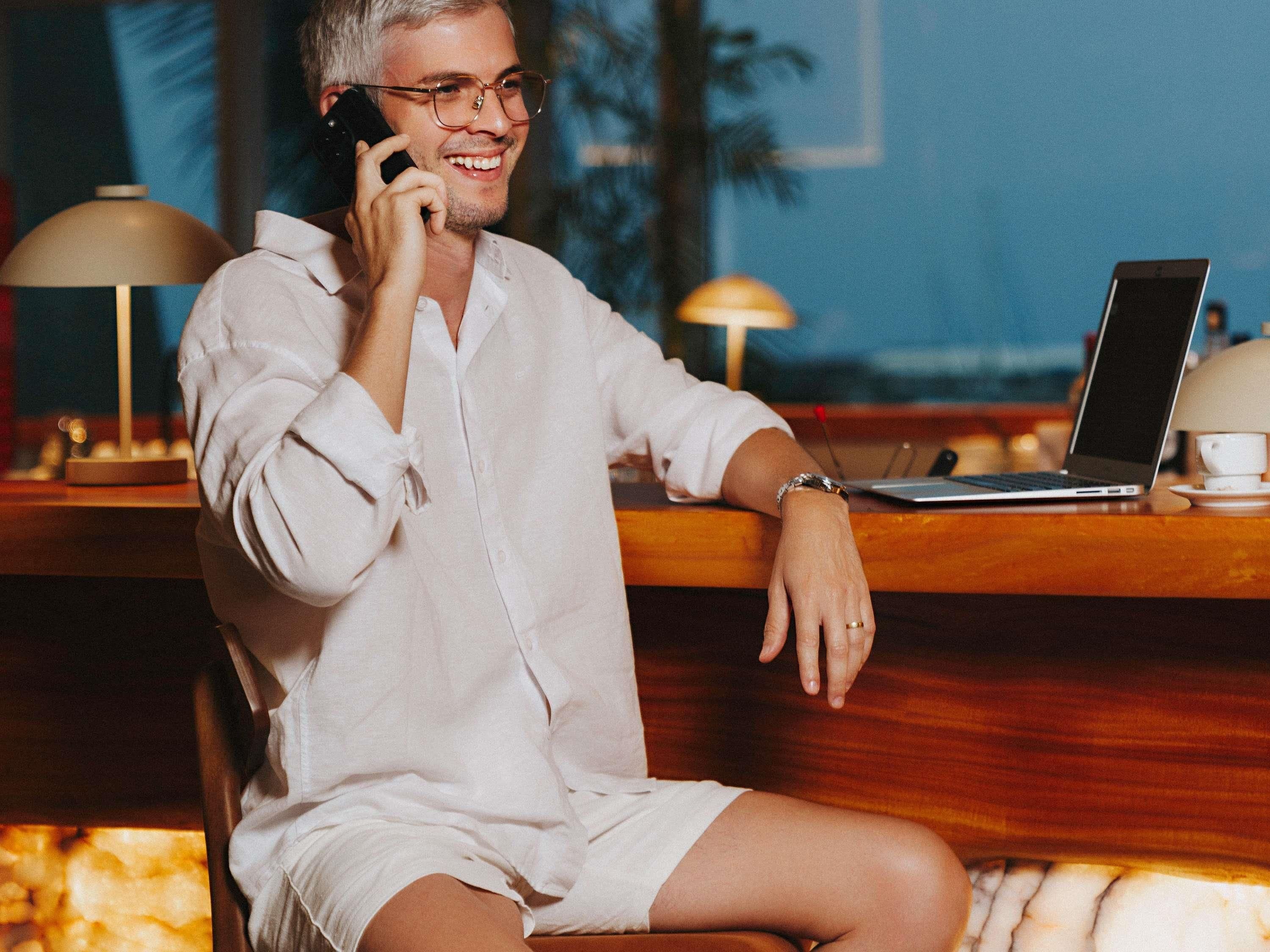 A person seated at a wooden table with a laptop, talking on a mobile phone.