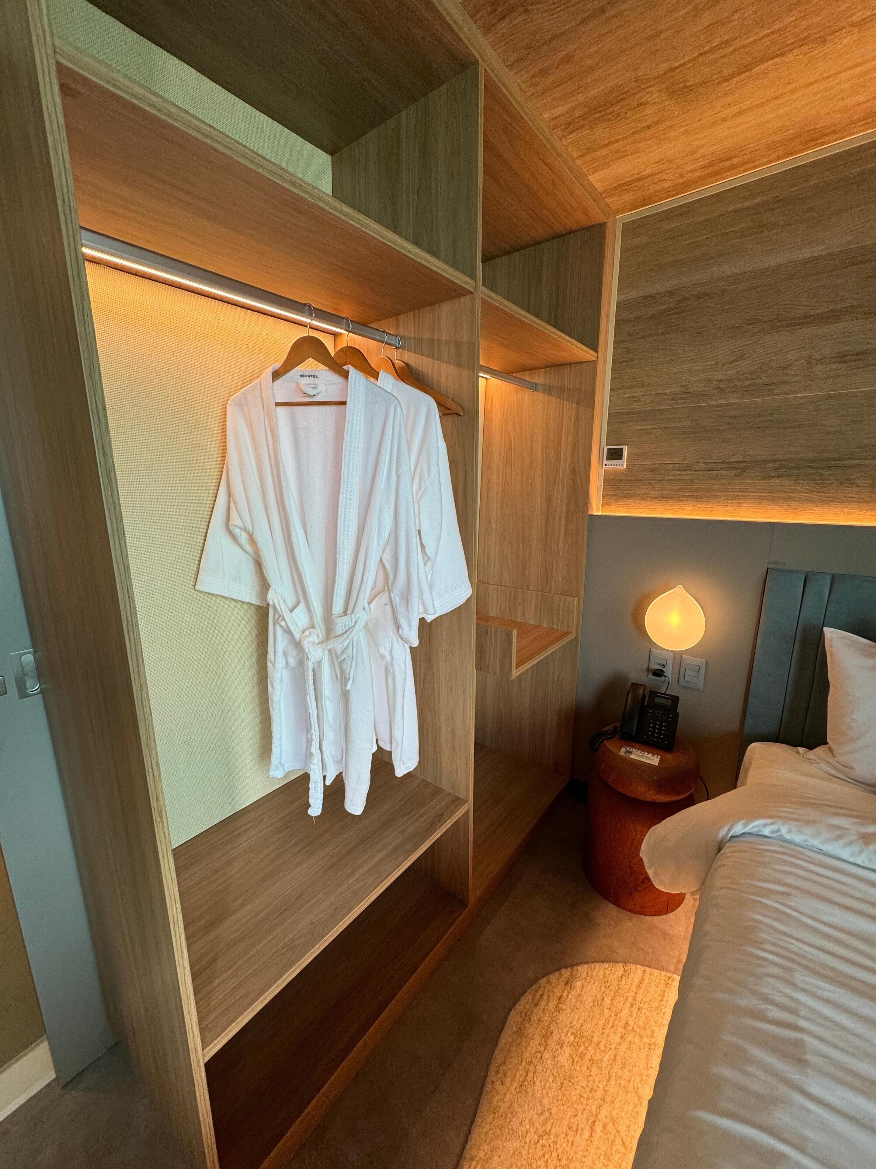 Habitación de hotel con armario de madera, cama con ropa de cama blanca, mesita de noche con lámpara y una bata colgada en el armario.
