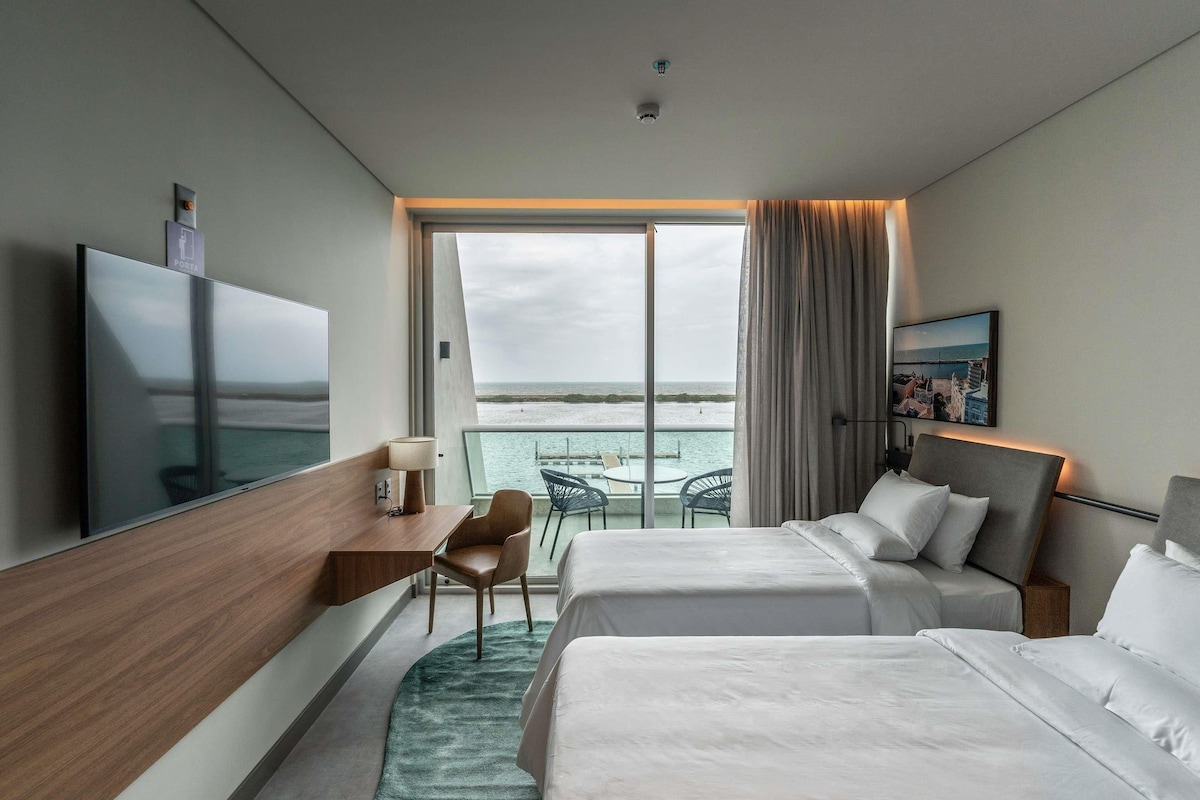 Una habitación de hotel moderna con dos camas, un escritorio, un televisor y un balcón con vista al mar.