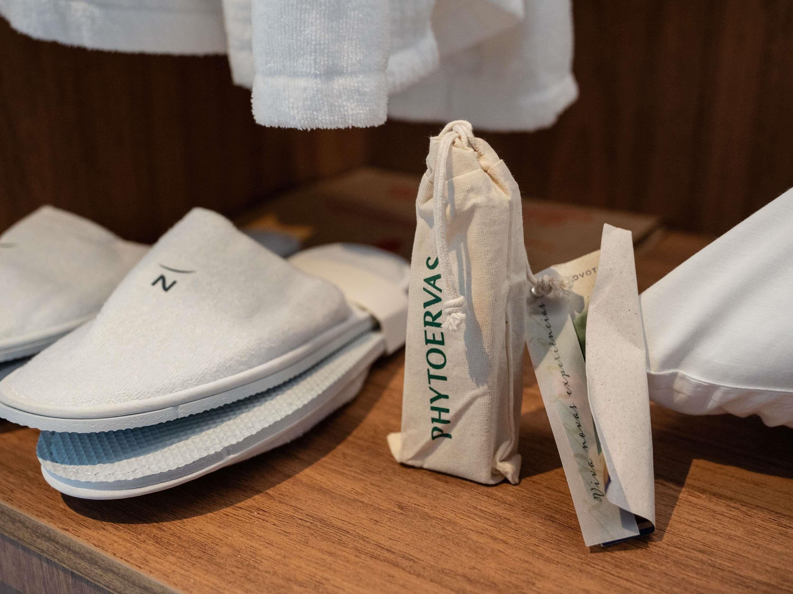 Uma superfície de madeira com um par de chinelos brancos, uma pequena bolsa branca com a inscrição 'PHYTOERVAS' e um tecido branco.