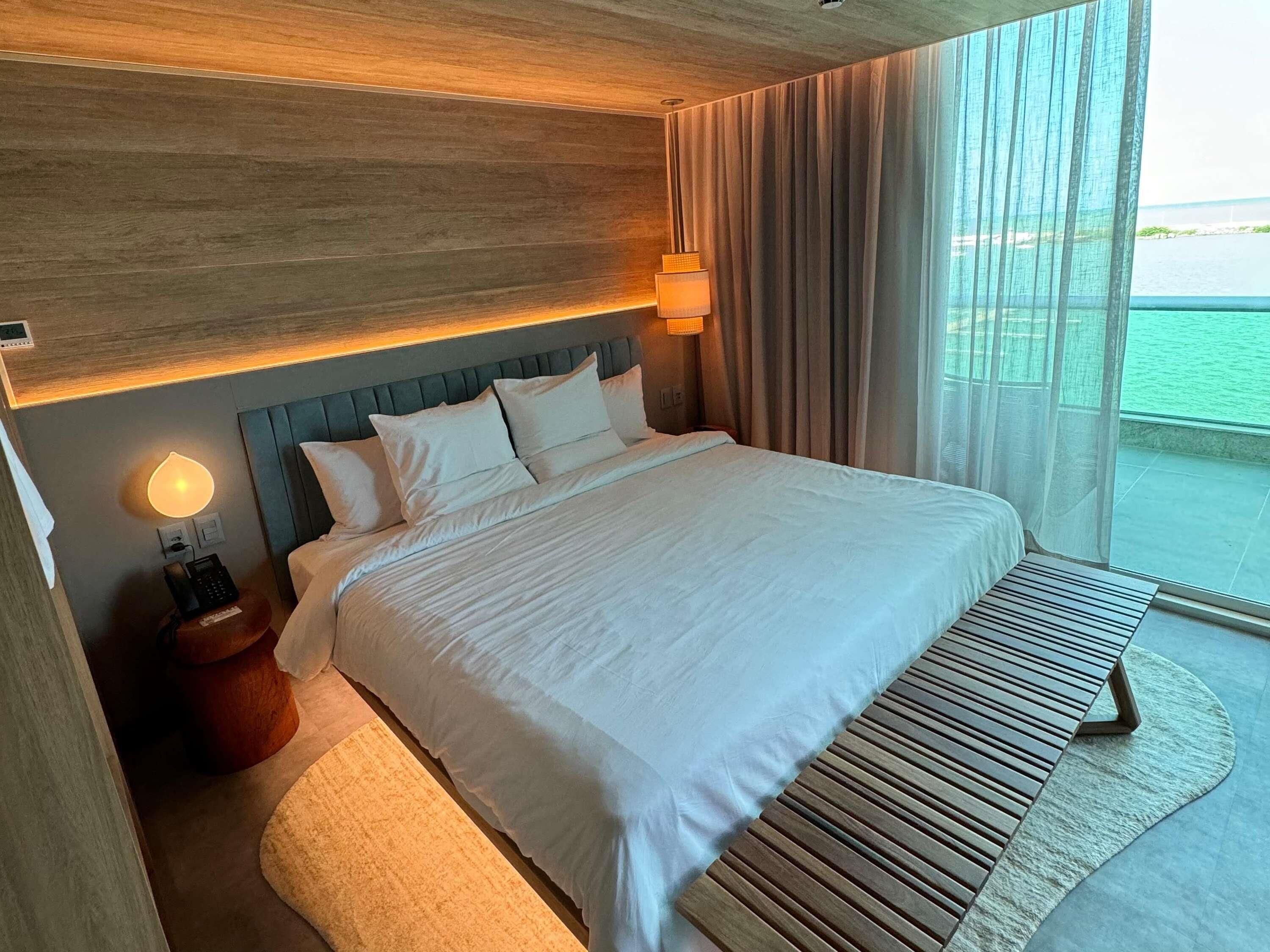 A hotel room with a large bed, a bedside table with a lamp, a phone, and a view of the sea.