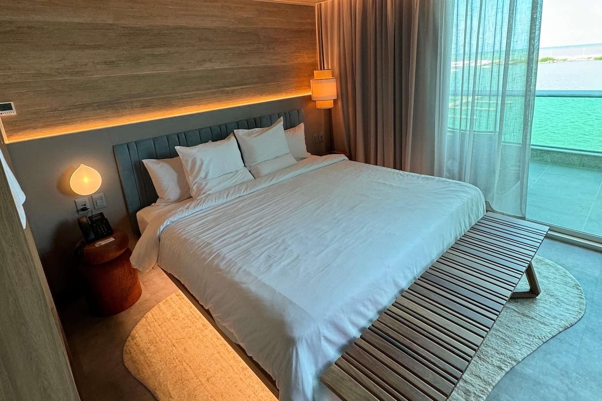 Ein Hotelzimmer mit einem großen Bett, einem Nachttisch mit Lampe, einem Telefon und Blick auf das Meer.