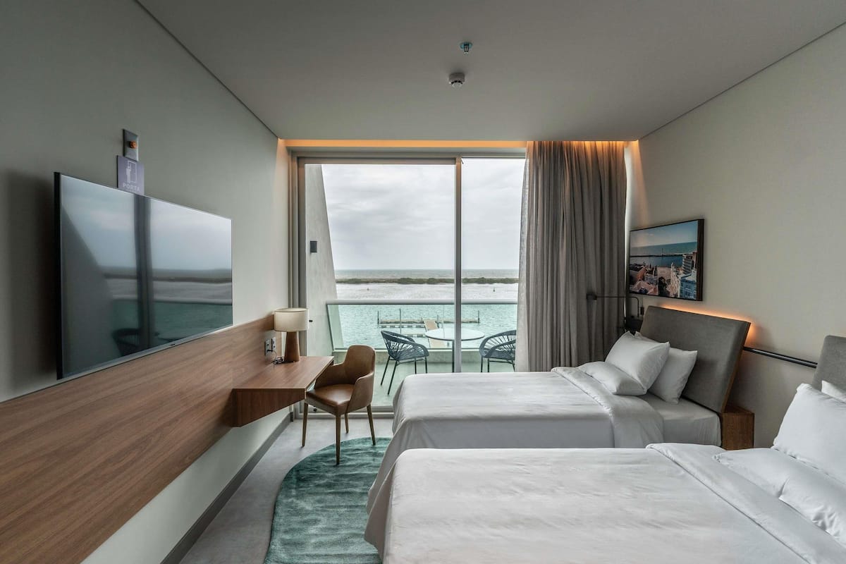 Ein modernes Hotelzimmer mit zwei Betten, einem Schreibtisch, einem Fernseher und einem Balkon mit Meerblick.