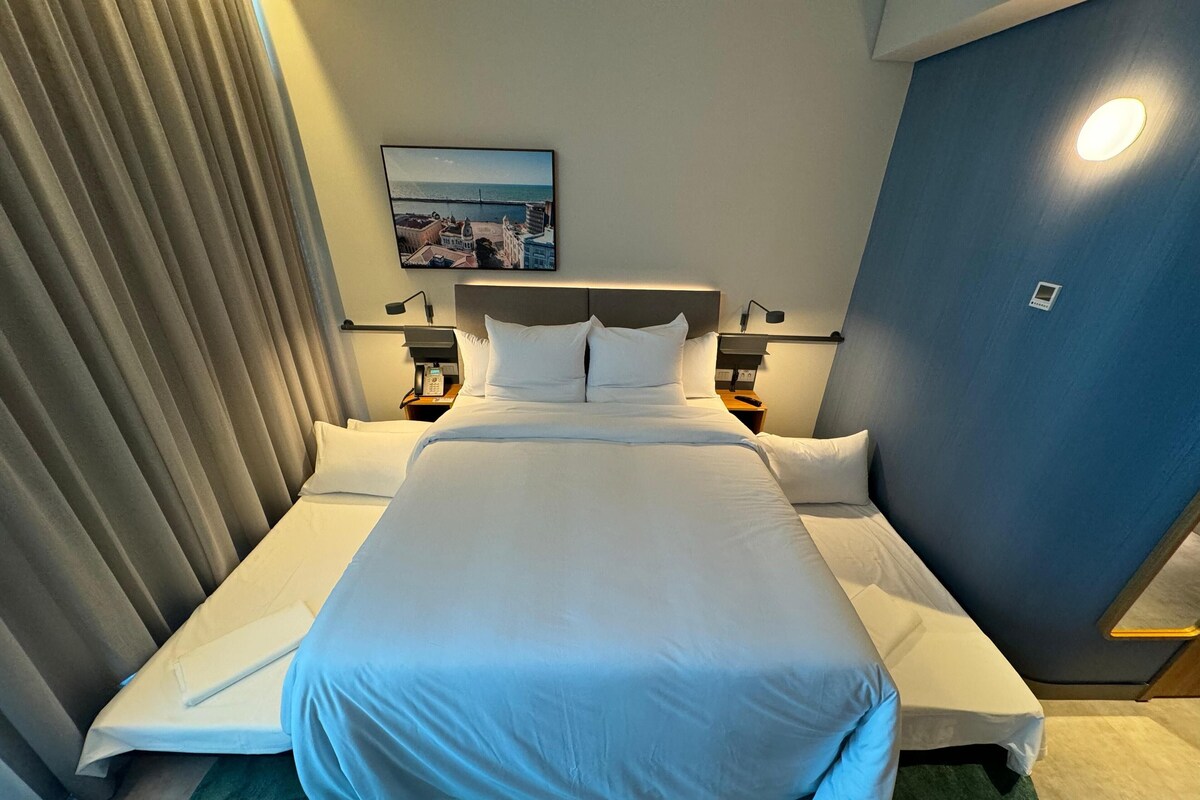 Una habitación de hotel con una cama grande, dos mesitas de noche, un teléfono en la pared y un cuadro con una vista costera.