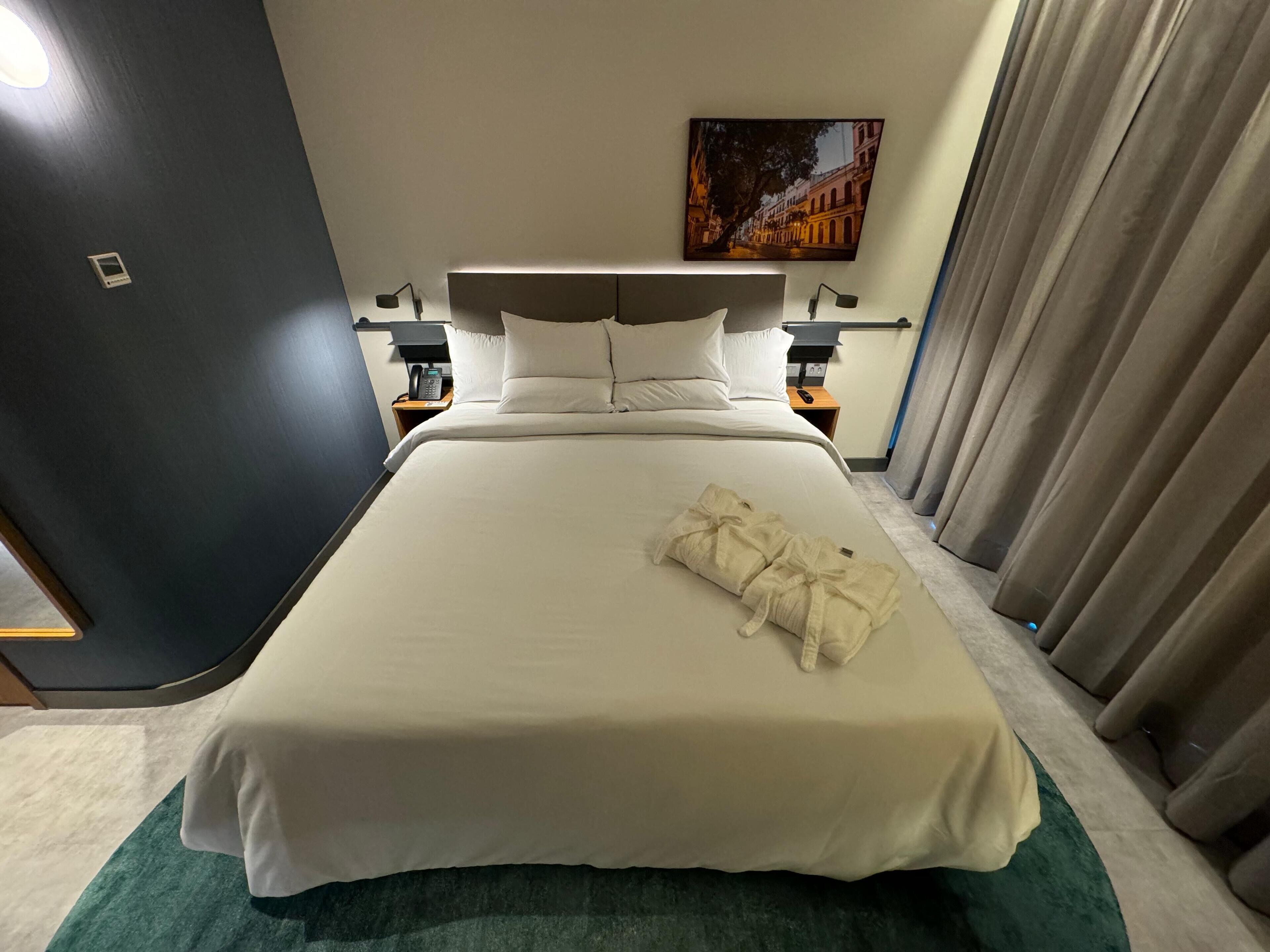 Una habitació d'hotel amb un llit gran, una tauleta de nit amb una làmpada, un quadre a la paret i una finestra amb cortines.