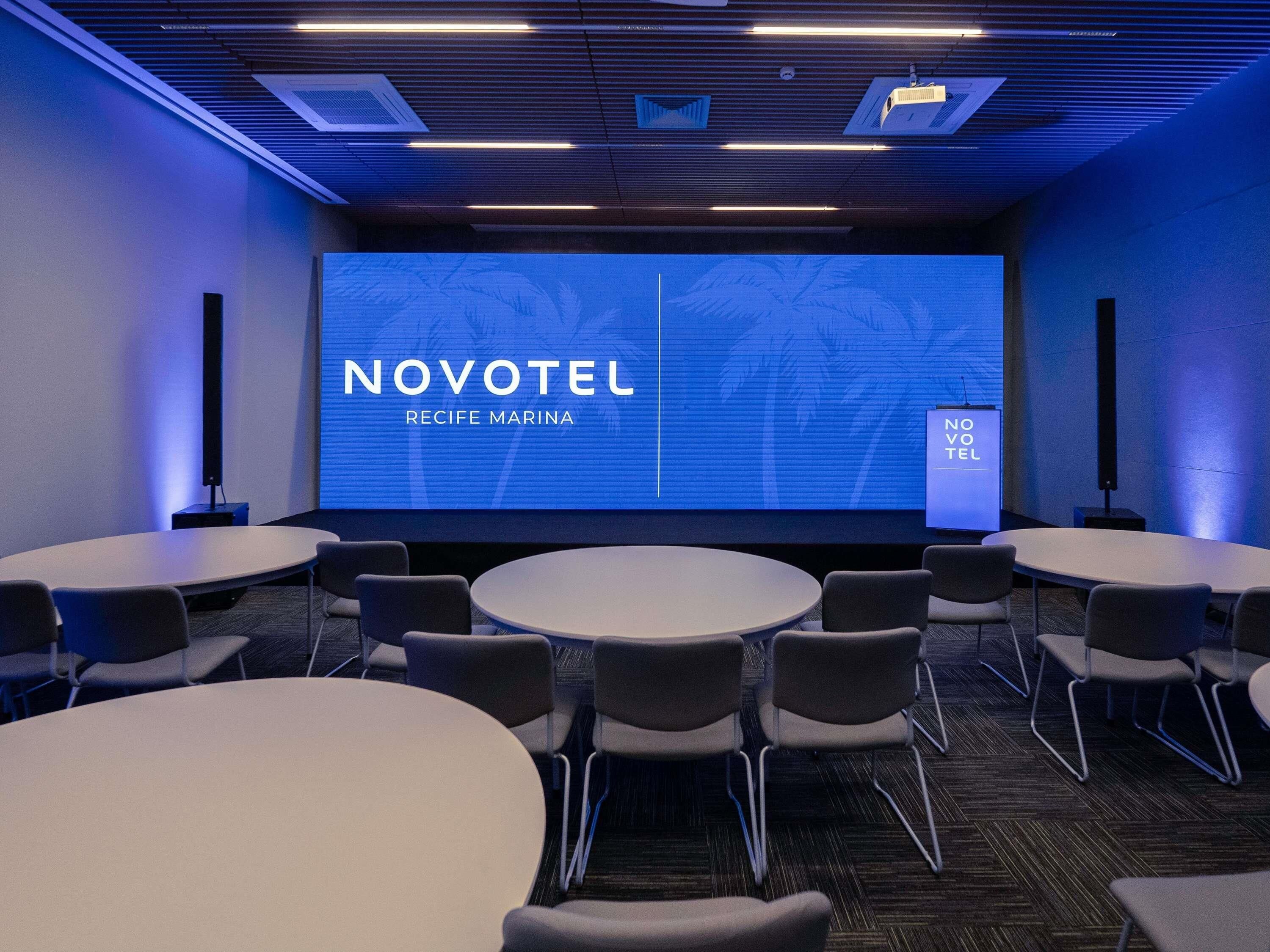 Een vergaderzaal met een groot scherm waarop 'NOVOTEL RECIFE MARINA' en 'NO VOTO TEL' staan. Er staan ronde tafels en stoelen klaar voor vergaderingen.