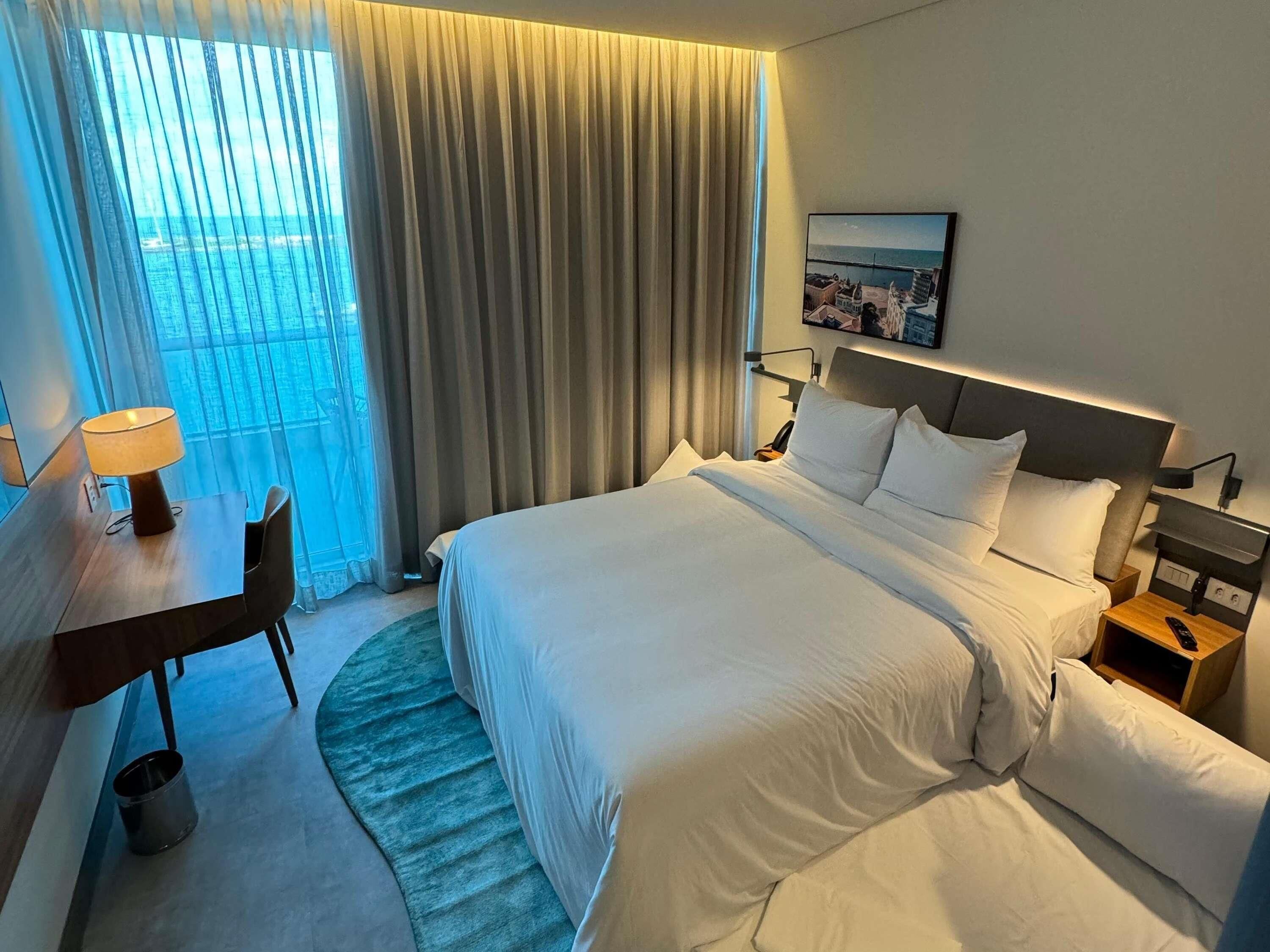 Ein Hotelzimmer mit Bett, Schreibtisch, Stuhl, Nachttisch, Lampe und Meerblick durch das Fenster.