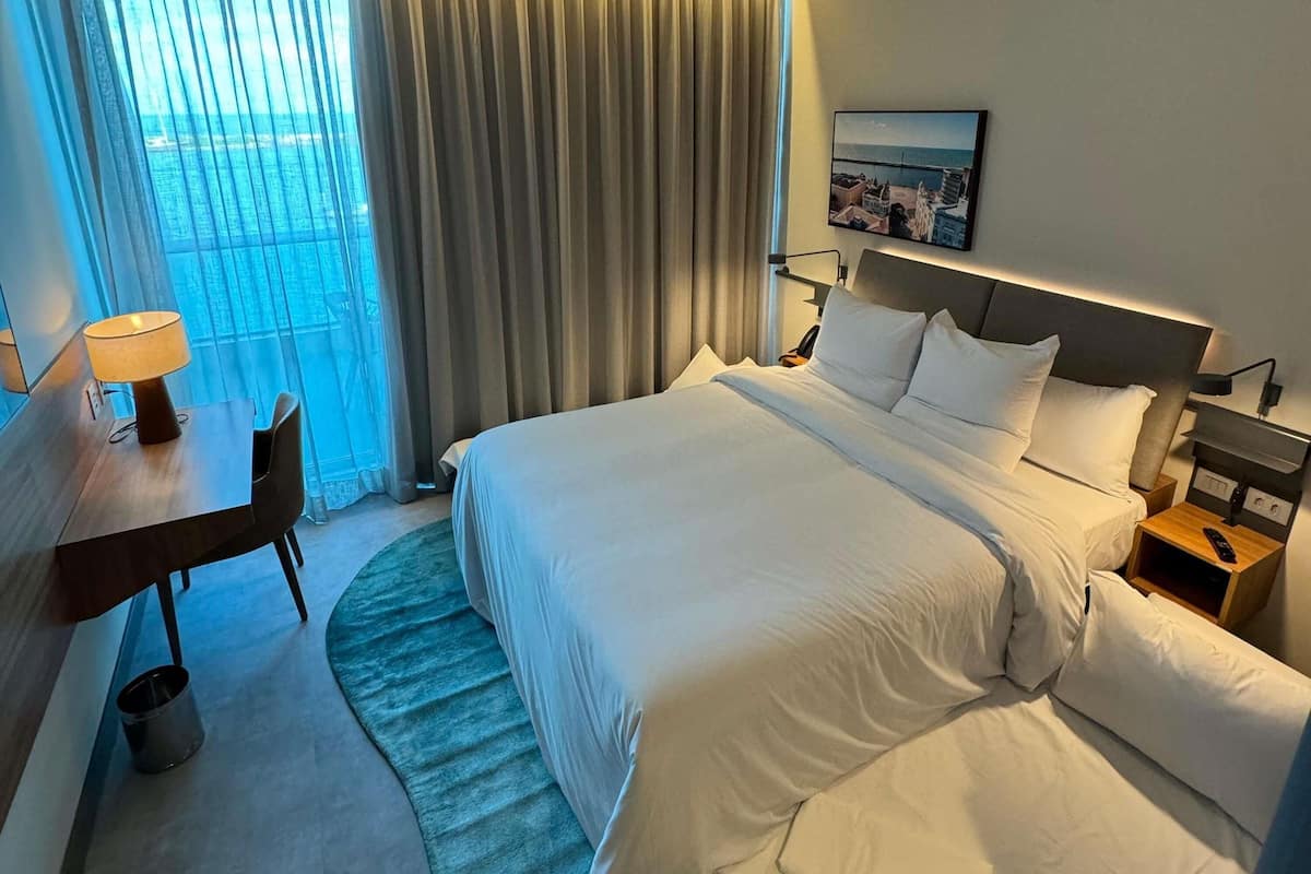 Ein Hotelzimmer mit Bett, Schreibtisch, Stuhl, Nachttisch, Lampe und Meerblick durch das Fenster.