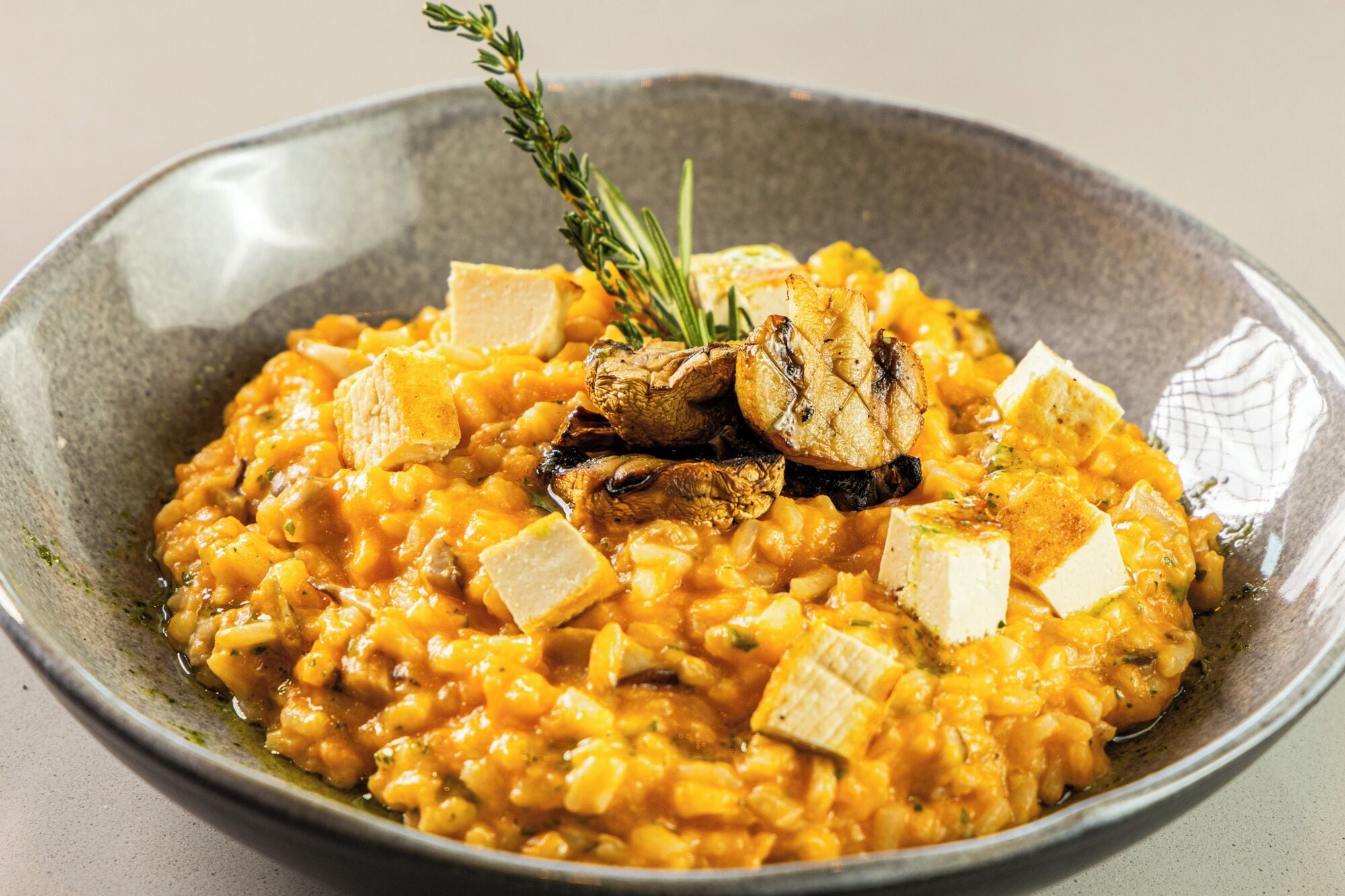 A bowl of creamy risotto with grilled mushrooms and cubes of cheese, garnished with a sprig of rosemary.