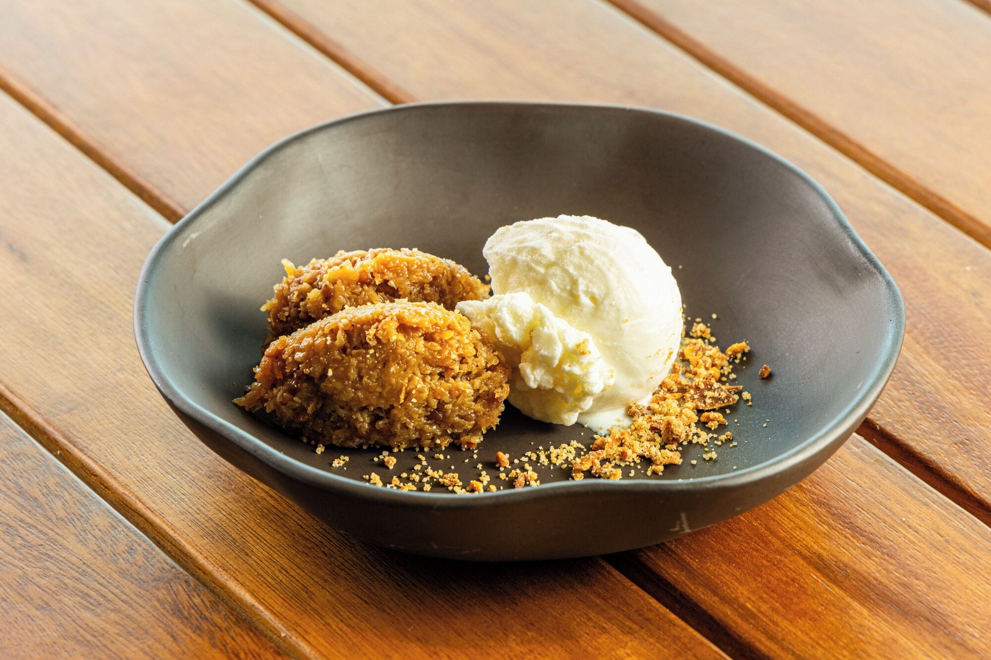 A dark bowl with two pieces of fried food and a scoop of ice cream on a wooden table.