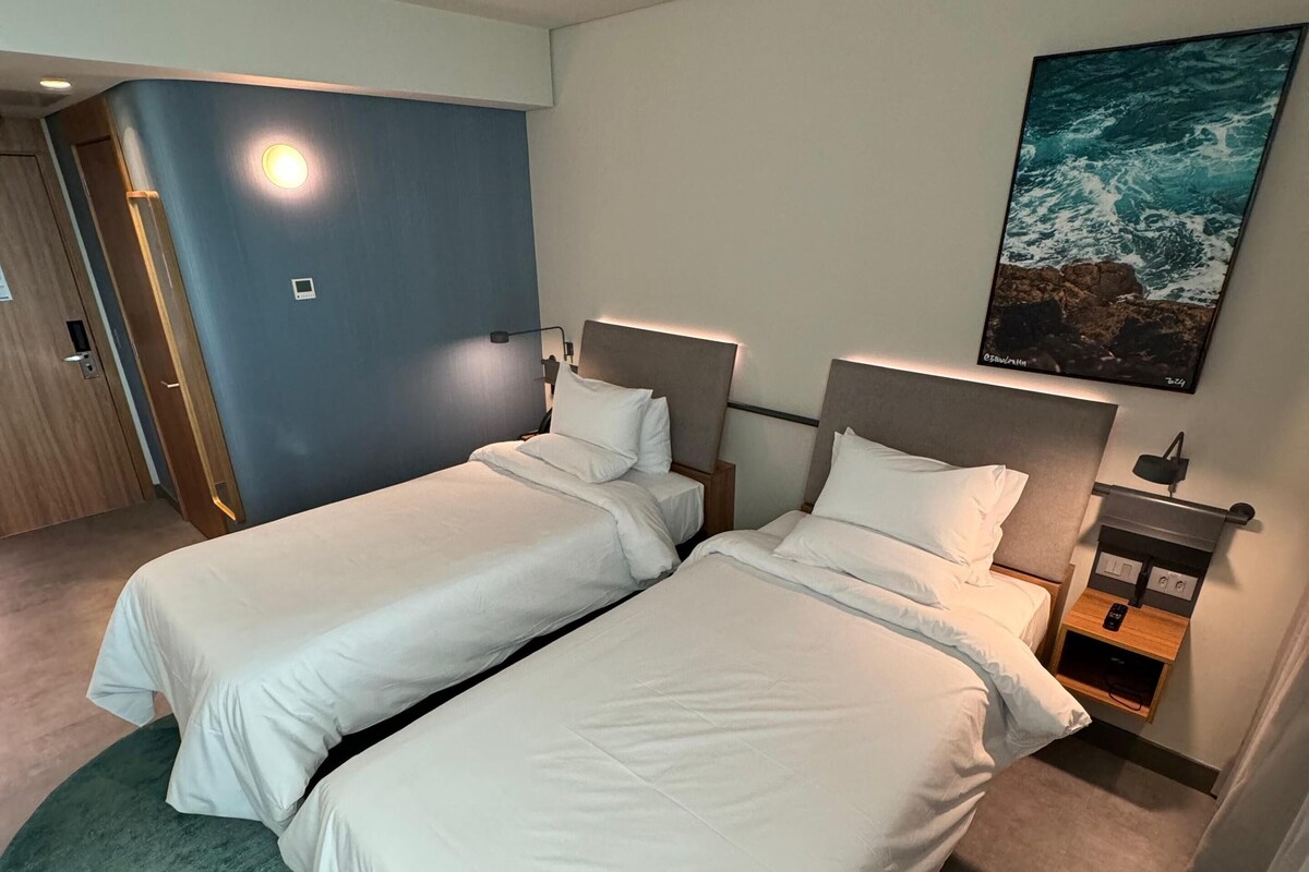 Una habitación de hotel con dos camas individuales, una mesita de noche con lámpara, un cuadro del océano y una puerta a la izquierda.